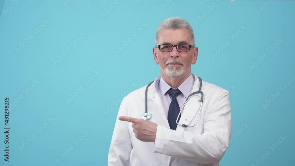 Man doctor in uniform and with a stethoscope pointing index finger to the side and looking after him in studio on blue background. Doctor points finger to the side.