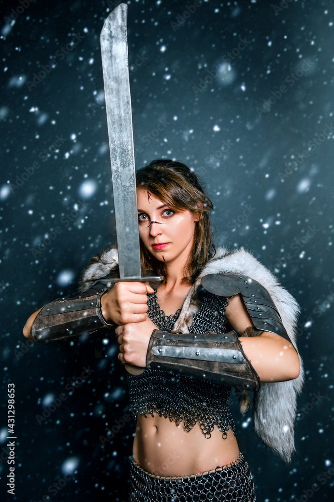 Portrait of a medieval woman warrior in chain mail and fur with a sword in her hands posing while standing in a snowstorm. Fantasy barbarian, viking.