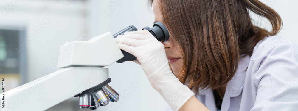 Obraz premium A young scientist woman in a laboratory coat looking through a microscope in a laboratory to do research and experiment. Scientist working in a laboratory. Education stock photo