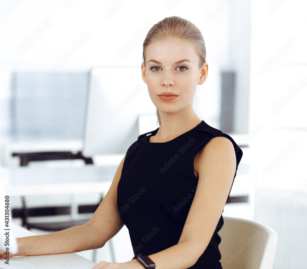 Young blond businesswoman working on computer while sitting at the desk in modern office. Business people concept. Secretary looks beautiful in black dress Young blond businesswoman working on computer while sitting at the desk in modern office. Business people concept. Secretary looks beautiful in black dress