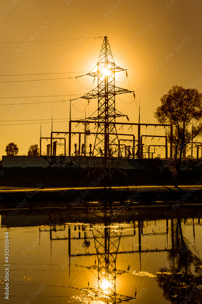 A power line on the shore of a pond against the background of a bright yellow sunset. Silhouette of an electric tower. Bright colors. Concept. Vertical orientation of the photo.