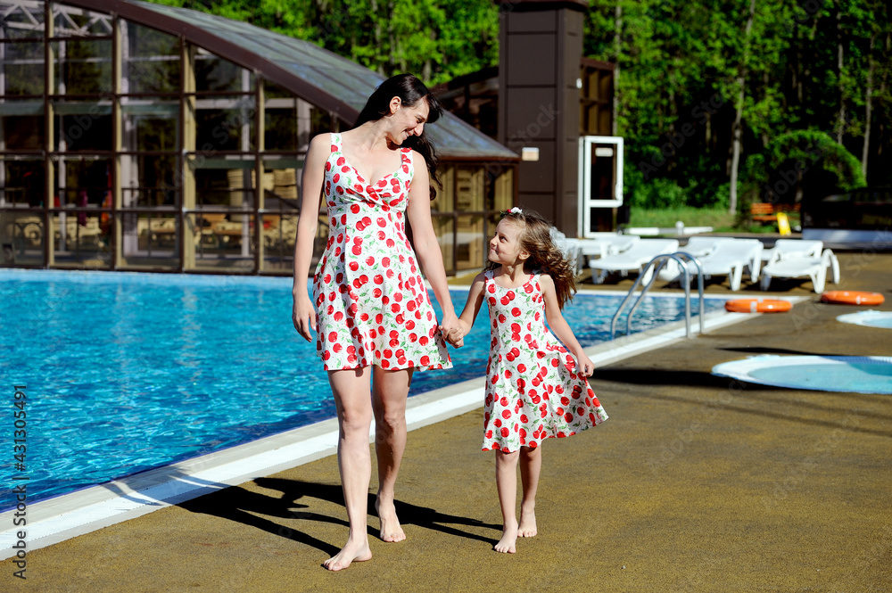 A pregnant woman and a little girl in beautiful matching dresses walk along the edge of the outdoor pool holding hands and looking at each other with a smile. Happy family. Horizontal orientation.