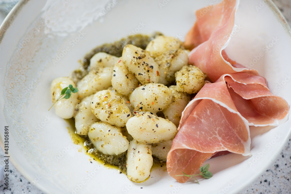 Italian gnocchi served with basil pesto sauce and prosciutto ham in a beige plate, selective focus, close-up, studio shot Italian gnocchi served with basil pesto sauce and prosciutto ham in a beige plate, selective focus, close-up, studio shot