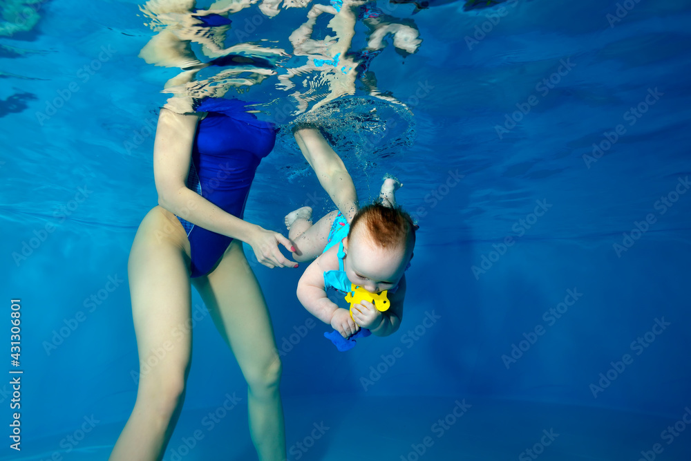A baby girl dives under the water in a children's pool with a yellow toy in her hands. A female instructor backs her up in the water. Children's swimming training. Concept. Horizontal orientation.