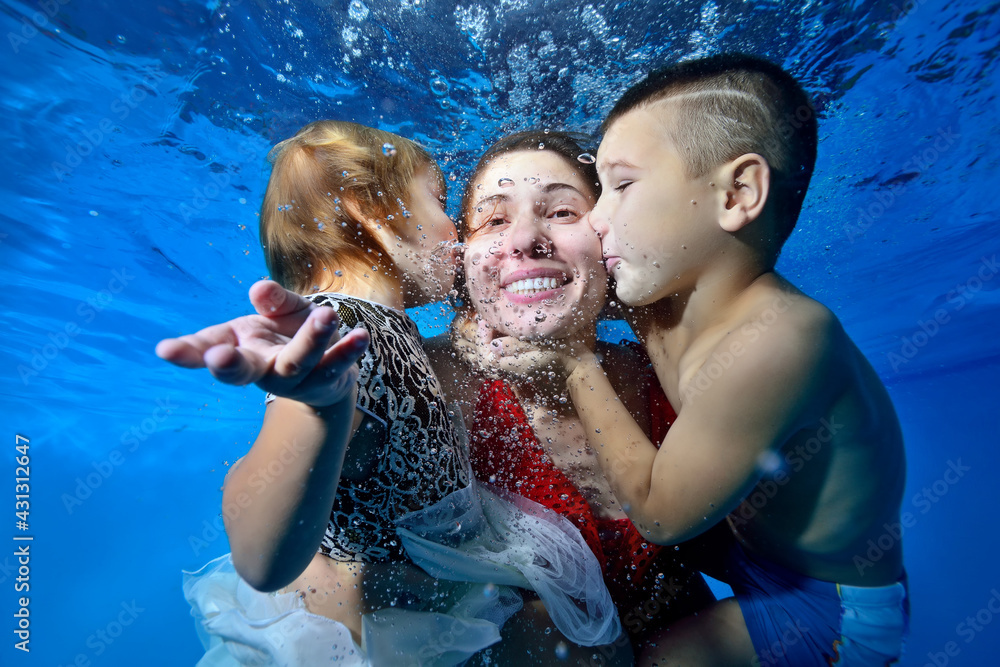 A young son and daughter kiss their mother from both sides while swimming underwater in the pool. They smile and play. The concept of a happy family. Portrait. Close-up. Horizontal view.
