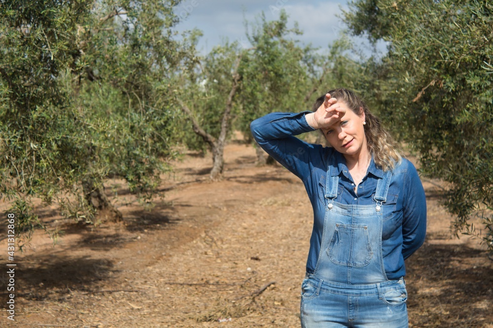 Blonde woman in jeans wiping sweat from her forehead with her arm in a field of organic olive trees on a sunny day Blonde woman in jeans wiping sweat from her forehead with her arm in a field of organic olive trees on a sunny day