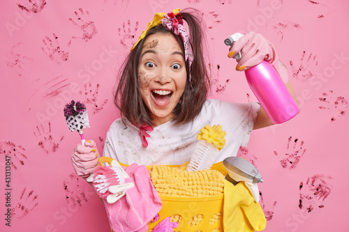 Funny positive housewife glad to finish house cleaning holds brush spray detergent stands dirty against pink background with dirty handprints. Professional cleaner goes to you. Domestic routines photo