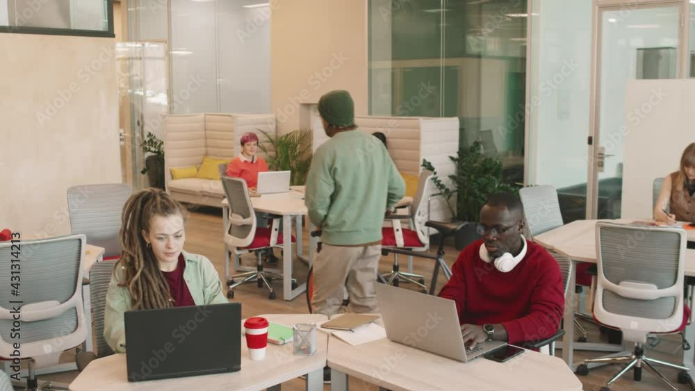 Tilt up slowmo shot of diverse young businesspeople working on laptops in open-plan office of creative start-up or coworking space. Bearded man in beanie hat parking his bicycle in office Tilt up slowmo shot of diverse young businesspeople working on laptops in open-plan office of creative start-up or coworking space. Bearded man in beanie hat parking his bicycle in office