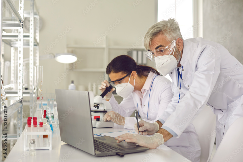 Microbiologist biotechnology researcher group in facial mask and protective gloves conducting experiment. Male supervisor making notes on laptop and female assistant analyzing sample using microscope Microbiologist biotechnology researcher group in facial mask and protective gloves conducting experiment. Male supervisor making notes on laptop and female assistant analyzing sample using microscope