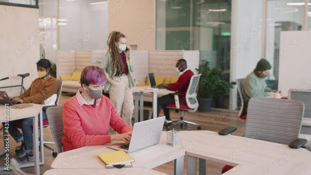 Slowmo PAN shot of diverse group of young businesswomen and businessmen in face masks working on laptops in open-plan office or coworking space Slowmo PAN shot of diverse group of young businesswomen and businessmen in face masks working on laptops in open-plan office or coworking space