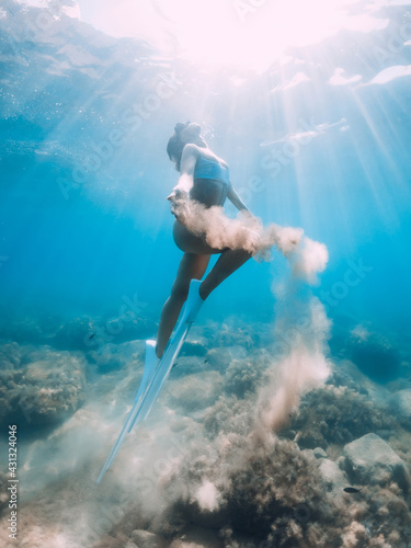 Freediver woman glides underwater with sand in hand. Free diver with fins posing in transparent sea