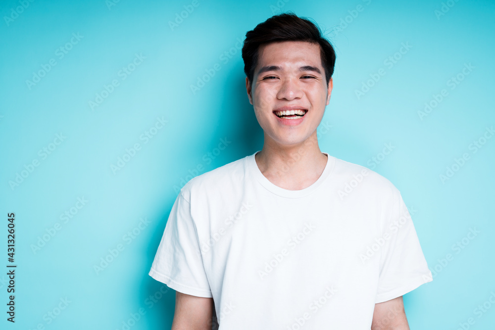 Happy young guy with asian appearance smiling and looking at camera while standing against blue background Happy young guy with asian appearance smiling and looking at camera while standing against blue background
