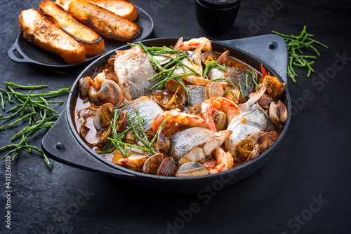 Modern style traditional Spanish seafood zarzuela de pescado with fish, king prawns and venus clams served with barbecue garlic bread in red sauce as close-up in design pot