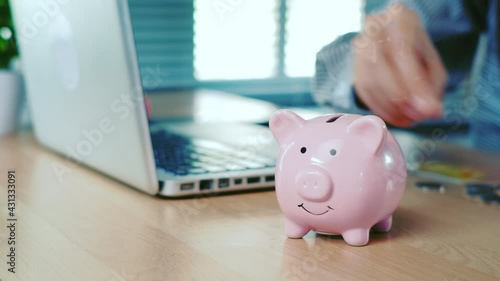 Business woman hand is putting a coin or money in a piggy bank on office desk. Saving money concept.
