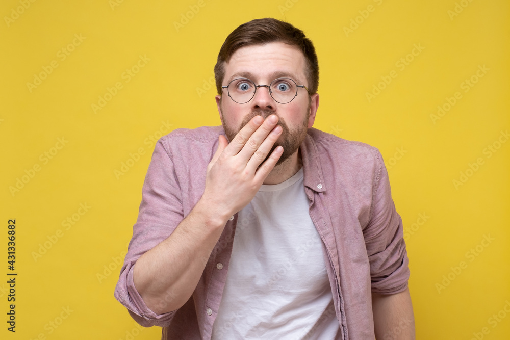 Frightened, amazed Caucasian man in round glasses looks shocked at the camera with hand over mouth. Yellow background. Frightened, amazed Caucasian man in round glasses looks shocked at the camera with hand over mouth. Yellow background.