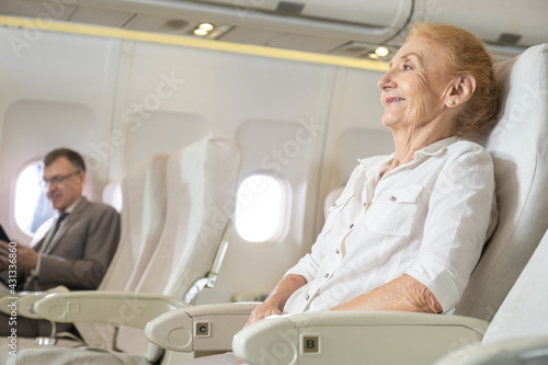 Happy senior Caucasian woman sitting or traveling in the plane and smiling with a blurred Caucasian businessman on background with copy space