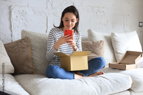 Beautiful young woman is holding cardboard box and unpacking smartphone sitting on sofa at home. photo