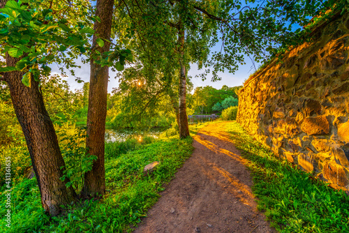 Alley going between the fortress and the river. The sun shines through the foliage of the trees. Beautiful nature. Priozersk, Russia. View from the road near the fortress wall.