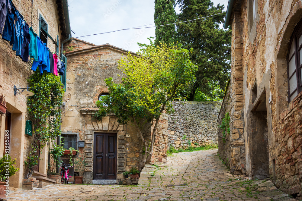 Fototapeta premium Narrow street in the old town of Volterra in a summer sunny day. Historic traditional houses and cobbled street with a tree growing on the side of an old house. Laundry is hanging from a balcony. Pisa
