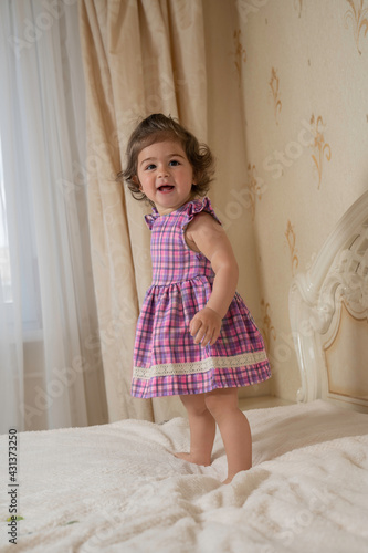 one-year-old girl with dark curly hair, brown big eyes stands in a dress on the bed