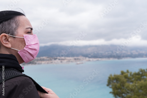 Short-haired woman dressed in black with a backpack and wearing a pink protective mask for the covid, stops on the road to look at the beautiful views towards the Mediterranean Sea. photo