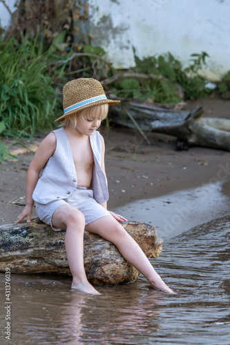 Fototapeta Naklejka Na Ścianę i Meble -  Sad and lonely little caucasian blond boy wearing straw hat sits by river. Fear of water. Mental Health Theme.