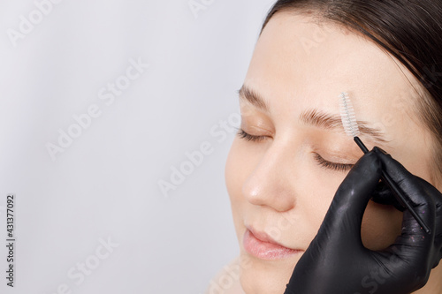 Master applies brow paste with a brush to eyebrows. Styling and lamination of eyebrows. Woman doing eyebrow permanent makeup correction. Gloved master combing patient’s eyebrows with a special brush.