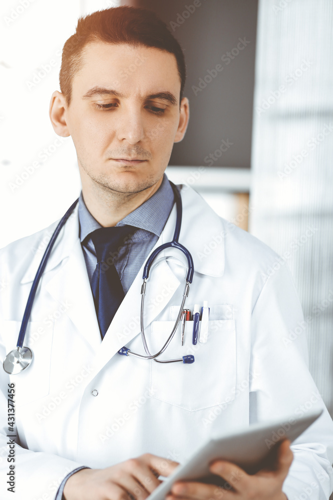 Friendly male doctor using tablet computer in clinic near his working place. Perfect medical service in hospital. Medicine and healthcare concept Friendly male doctor using tablet computer in clinic near his working place. Perfect medical service in hospital. Medicine and healthcare concept
