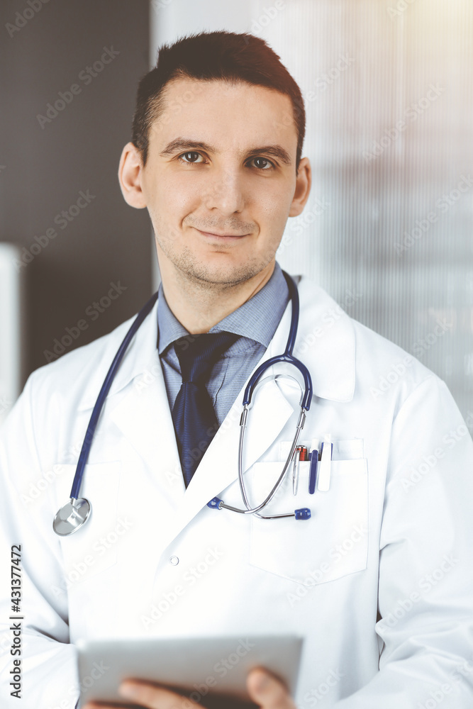 Friendly male doctor using tablet computer in clinic near his working place. Perfect medical service in hospital. Medicine and healthcare concept Friendly male doctor using tablet computer in clinic near his working place. Perfect medical service in hospital. Medicine and healthcare concept