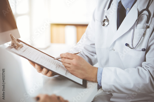 Unknown male doctor and patient woman discussing current health examination while sitting in sunny clinic and using clipboard. Perfect medical service in hospital