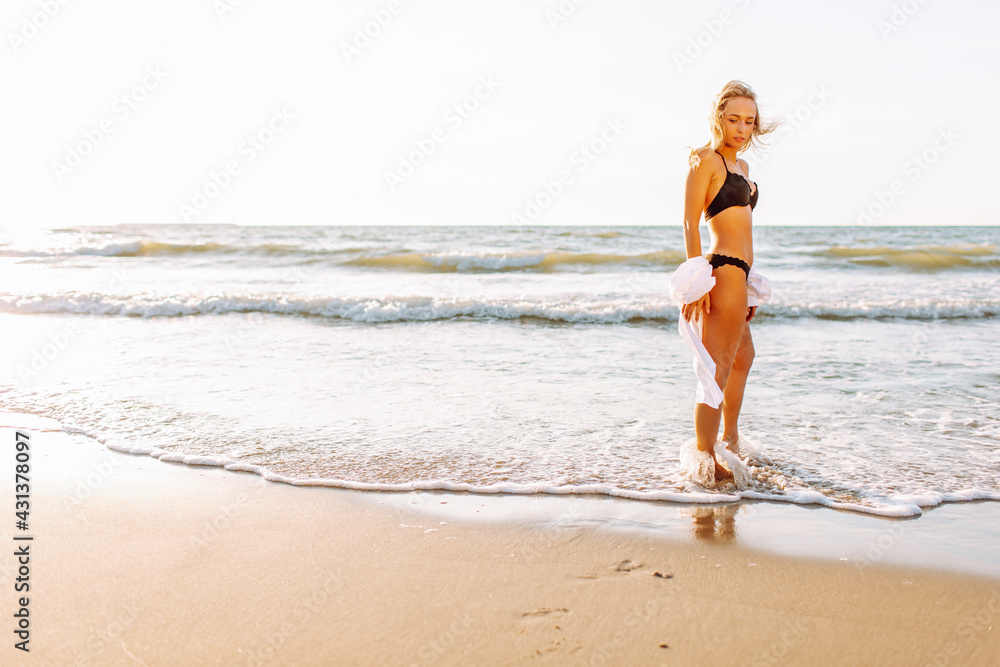 Fototapeta premium Beautiful blonde slim woman wearing black bikini walk on beach of sea or ocean against the sun. Youth, vacation, travel, fashion underwear