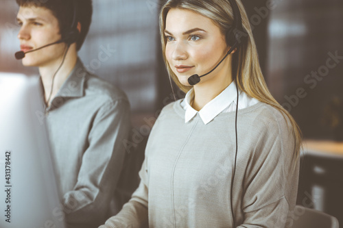 Diverse people group in sunny call center. Blonde business woman talking by headset while sitting in modern office