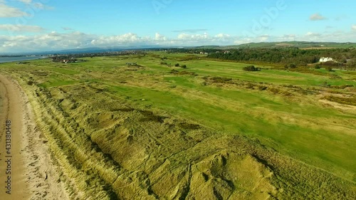 Aerial Panning High Above The Lush Greens Of The Royal Troon Golf Club, With Blue Skies And Exclusive Homes Nestled In The Trees photo