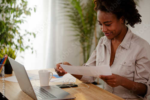 Smiling Afro American businesswoman working from home, drinking a coffee and holding some papers.