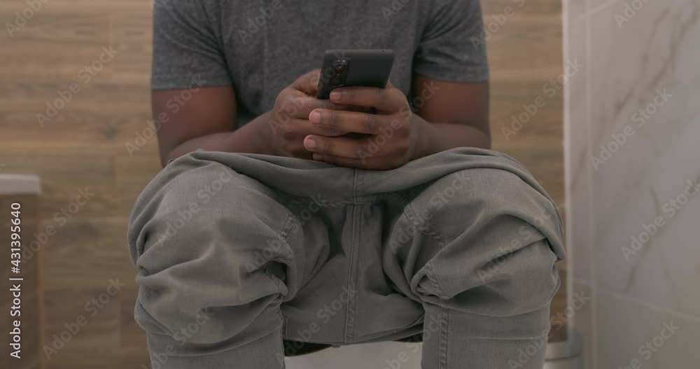 Cropped African man using smart phone while sitting on toilet in bathroom. Chatting in restroom Cropped African man using smart phone while sitting on toilet in bathroom. Chatting in restroom