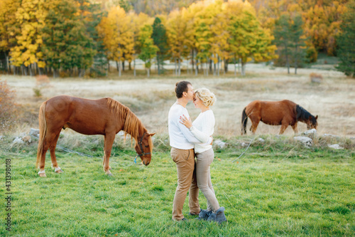 Man hugs and kisses woman on the forehead against the background of grazing horses in the autumn forest