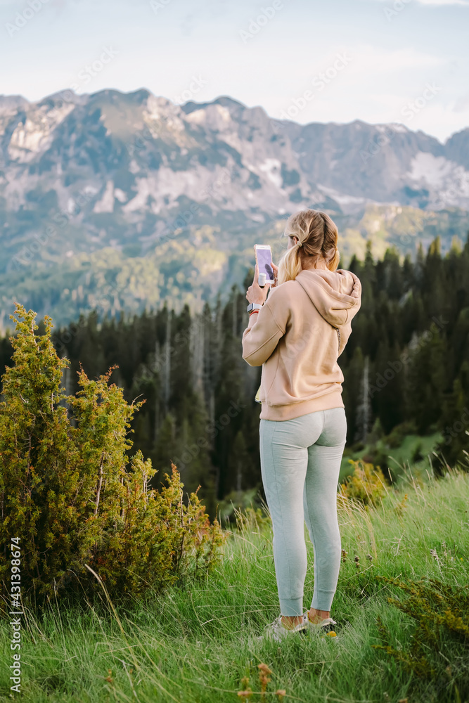 Hipster girl is shooting video of beautiful landscape on smartphone. Mountain scenery background with copy space. Female tourist is taking photo with mobile phone camera. 