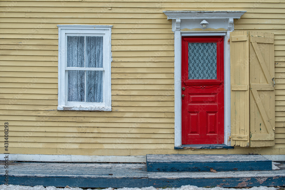Fototapeta premium The exterior wall of a yellow wood clapboard heritage house. The door of the building is bright red with a small glass window. There's a lace curtain in the double hung window and an iron patio bench.