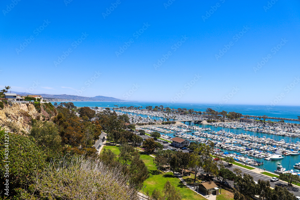 a stunning shot of the boats and yacht docked and sailing Dana Point Harbor with deep blue ocean water, lush green plants and trees on the hillside and blue sky in Dana Point California	 - obrazy, fototapety, plakaty 