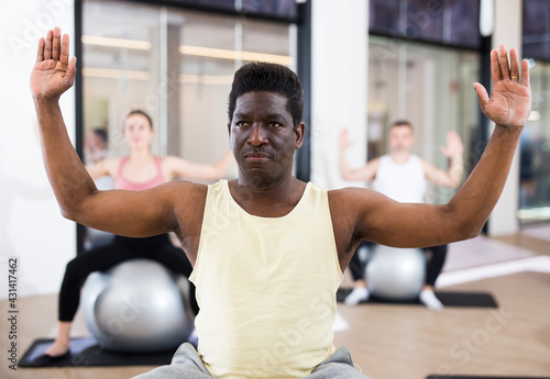 Confident young adult man doing exercises with pilates ball during group training at gym