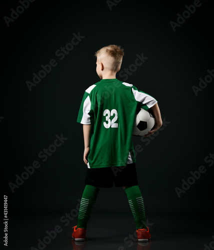 Young soccer player  football school student  teenager boy in red white striped uniform stands back to camera holding ball over black background. Back view