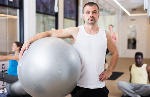 Portrait of sporty man standing with fitball during pilates group workout at gym. Healthy lifestyle concept
