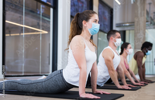 Young woman in protective face mask performing set of pilates exercises with group in fitness studio. New life reality in pandemic..