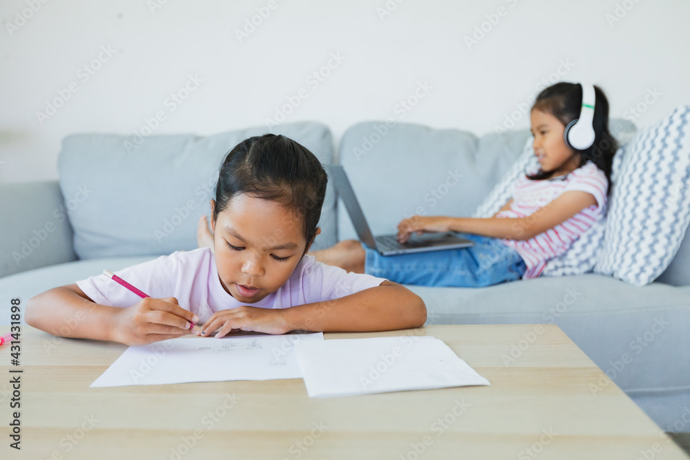 Asian child girl doing worksheet with her sister sitting on sofa and having video conference chat with teacher and class group. Children are homeschooling during quarantine due to COVID-19 outbreak. Asian child girl doing worksheet with her sister sitting on sofa and having video conference chat with teacher and class group. Children are homeschooling during quarantine due to COVID-19 outbreak.