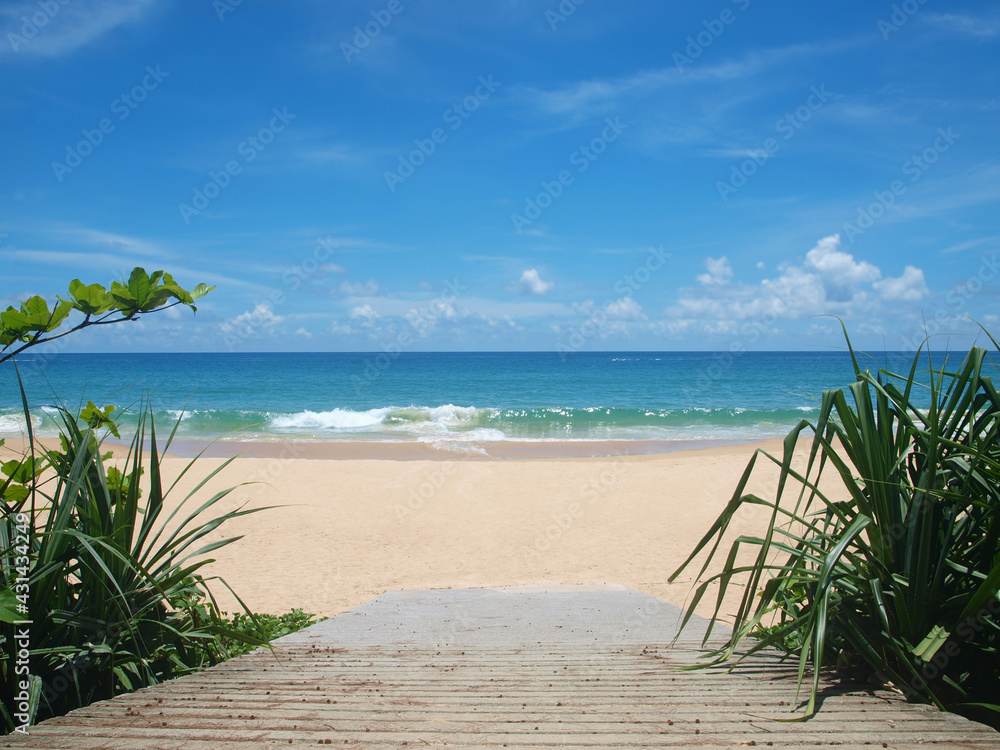 Beautiful view of tropical beach. Road extending to the sandy empty beach. Green trees on the side of cement path. Clouds on the sky. Blue sea water, moving watercraft on the surface. Bright colors