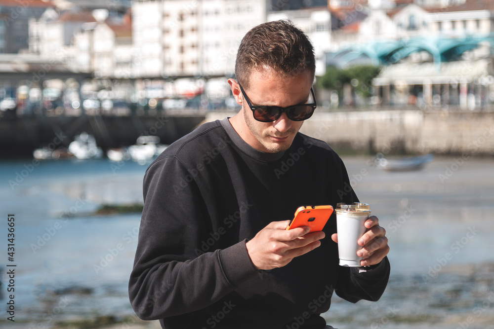 Young man with sunglasses drinking takeaway coffee and using mobile phone sitting on the street with yacht harbor in the background. Travel concept Young man with sunglasses drinking takeaway coffee and using mobile phone sitting on the street with yacht harbor in the background. Travel concept
