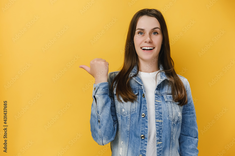 Positive excited young brunette woman 20s points thumb, demonstrates copy space on blank yellow studio background wall, has happy friendly expression, wears blue denim jacket. People emotion concept Positive excited young brunette woman 20s points thumb, demonstrates copy space on blank yellow studio background wall, has happy friendly expression, wears blue denim jacket. People emotion concept