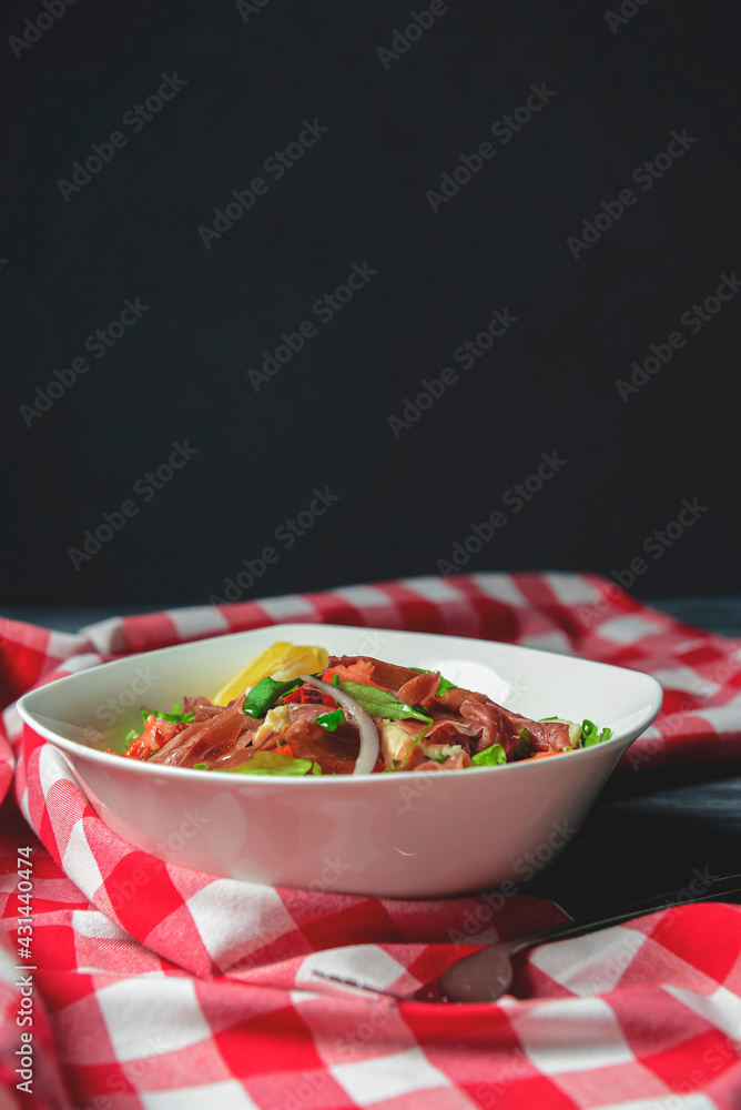 Prosciutto and arugula salad in a white bowl over wooden background. Red plaid tablecloth.