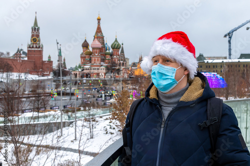 Portrait of caucasian man wearing blue protective face mask and red Santa hat. Blurred Saint Basil's Cathedral and Moscow Kremlin in the background. Theme of Christmas in Russia during coronavirus.