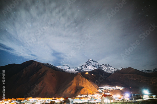 Stepantsminda, Georgia. Real Night Starry Sky With Glowing Stars Over Peak Of Mount Kazbek Covered With Snow. Famous Gergeti Church And Countryside Houses In Lightning. Georgian Landscape.